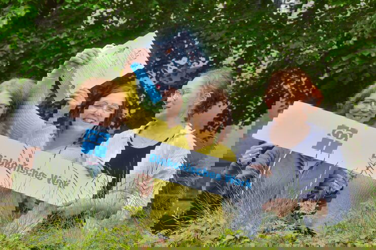 Ein Foto von drei weiblich gelesenen Personen, die in die Kamera lächeln. Sie halten ein weißes Banner mit blauer Schrift "Diakonisches Werk". Die mittlere Person hält eine Urkunde. Sie stehen im Grünen. 