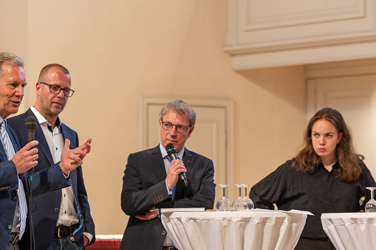 Drei männlich gelesene Personen und zwei weibliche stehen zusammen an weißen Tischen. Einer hält ein Mikro in der Hand.