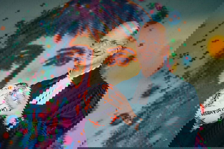 Eine als Mann lesbare Person im Talar mit einem Fußball vor einem Jesus-Graffito