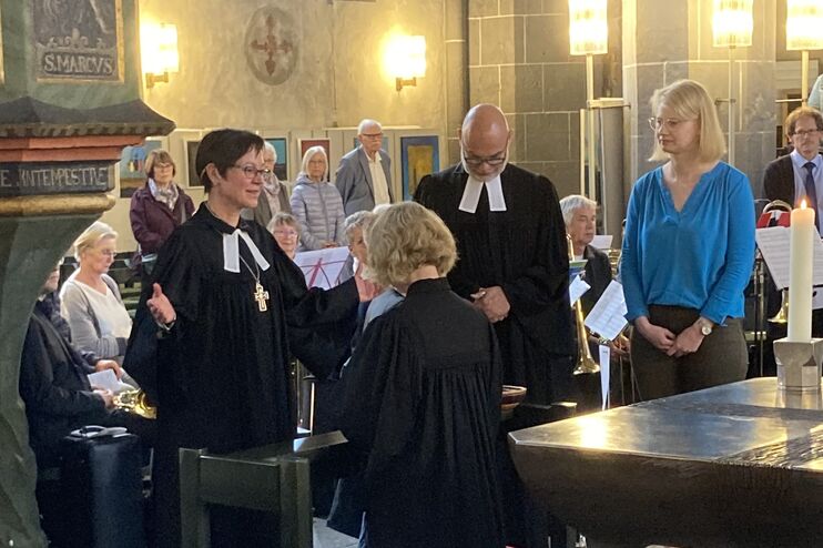 Eine weiblich gelesene Person im Talar steht, die Hände nach oben offen zur Seite ausgestreckt, vor einer knienden Person. Um sie stehen weitere Personen, eine mit Talar, in einem Kirchenraum.