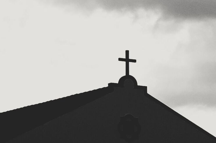 Gegen einen grauen Himmel zeichnet sich auf einem Spitzdach ein Kreuz ab.
