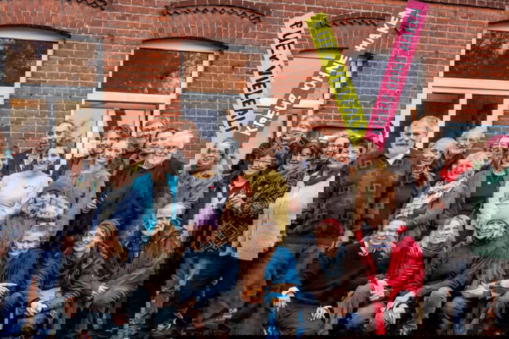 eine Gruppe von Menschen vor einem Backsteingebäude. Im Hintergrund gelb-magentafarbene Banner mit der Aufschrift "Für Vielfalt", "Kreuz ohne Haken"