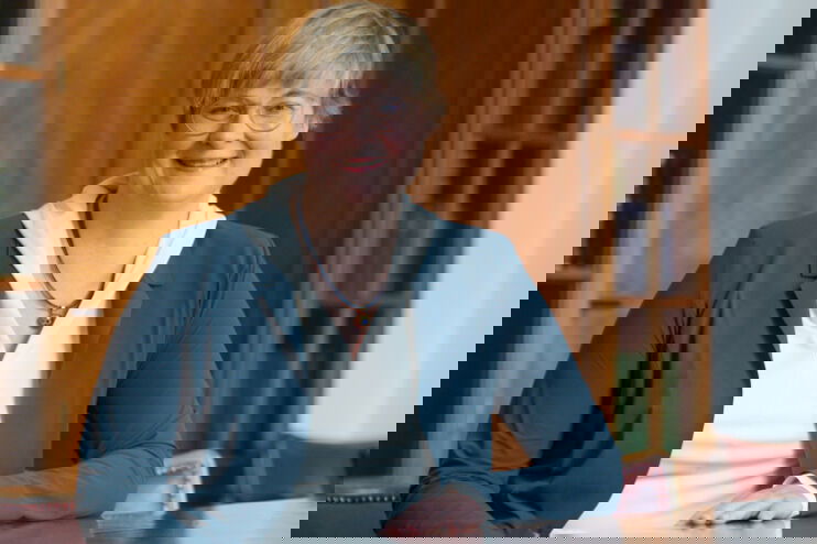 eine als Frau gelesene Person sitzt an einem Besprechungstisch in einem Büro. Hinter ist ist ein Holzschrank mit Büchern.