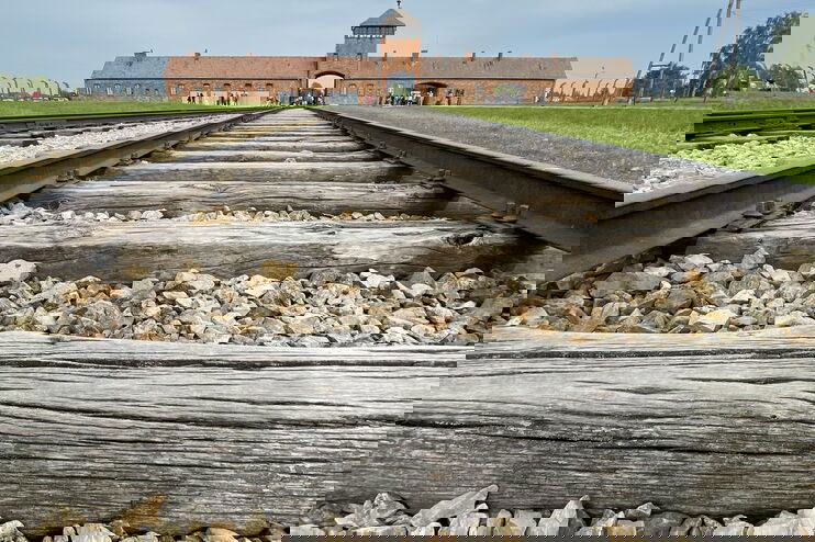 Blick aus der Froschperspektive über Gleise, die zu einem großen Gebäude führen - dem KZ Auschwitz.