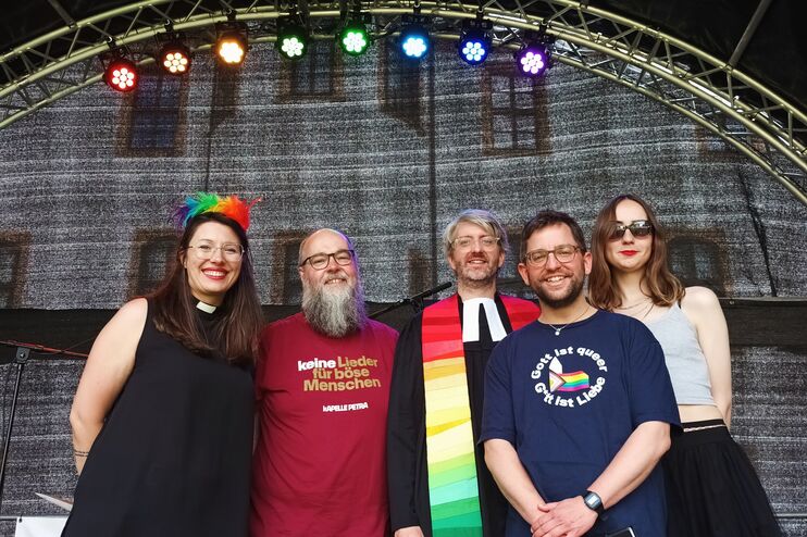 Fünf Menschen auf einer Bühne mit bunten Scheinwerfern im Hintergrund. Manche tragen regenbogen-bunte Accessoires.