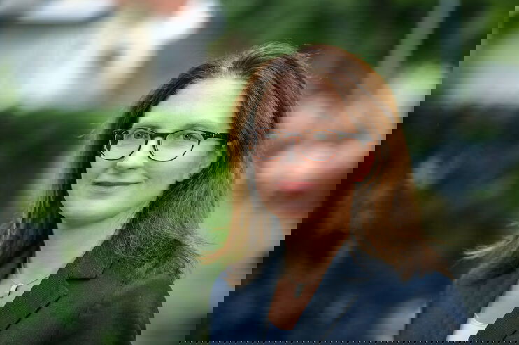 Eine weiblich gelesene Person mit Brille und mittellangen, braunen Haaren und Brille in blauem Jackett steht draußen vor einer grünen Hecke.