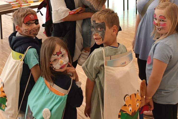 Vier Kinder stehen neben zwei Erwachsenen, einer hat auf ein Kind auf dem Arm. Die Kinder haben geschminkte Gesichter (Pirat, Schmetterling, Fledermaus) und tragen Stoffbeutel.