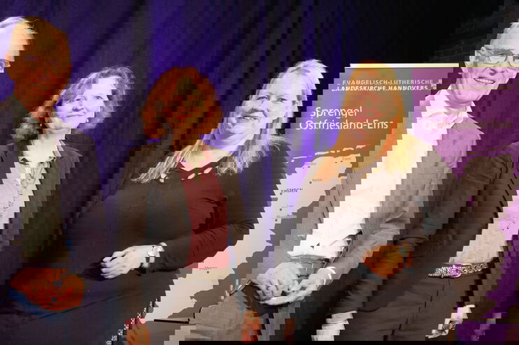 Eine als Mann und zwei als Frauen lesbare Personen posieren vor einem Aufsteller des Spremgels Ostfriesland-Ems