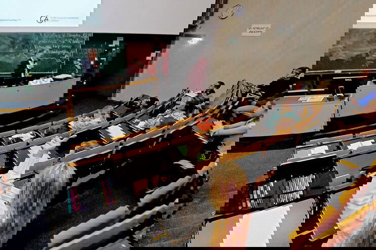 Kinder hören in einem Hörsaal einem Professor zu. Eine als Mädchen lesbare Person meldet sich.