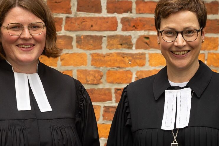 Zwei Frauen in einem Talar stehen vor einer Backsteinmauer und lächeln