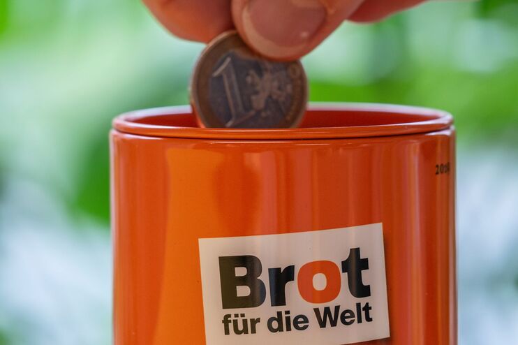 Orangfarbene Spendendose mit dem Logo von Brot für die Welt, eine Hand steckt eine Euromünze hinein.
