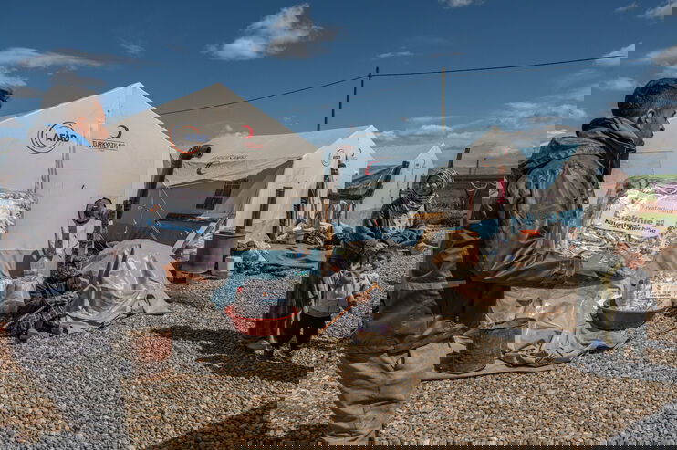 Eine männlich gelesene Person trägt ein Paket, auf dem "Diakonie Katastrophenhilfe" steht, zu einer weiblich gelesenen Person mit Kind. Das Umfeld wirkt wie in einem Flüchtlingslager: Zelte, Kiesboden.