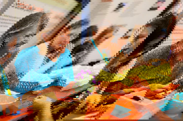 Weiblich gelesene Personen unterschiedlichen Alters sitzen an einem langen Tisch und knüpft Perlenarmbänder, sie tragen den Kirchentagsschal aus Nürnberg, manche unterhalten sich und lachen