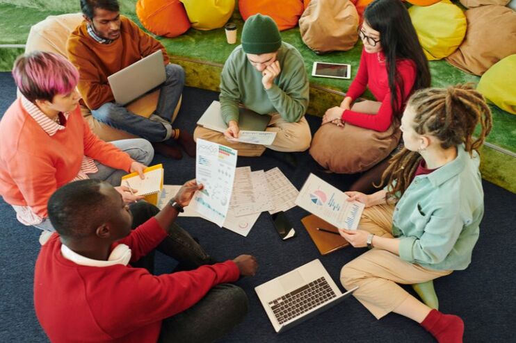 Mehrere junge Menschen unterschiedlicher Hautfarbe sitzen zusammen auf dem Boden, ein Laptop und mehrere Zettel zwischen sich.