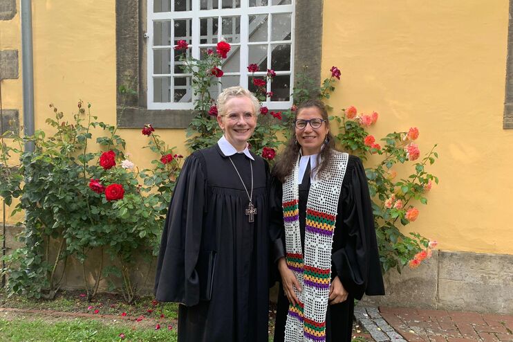Zwei weiblich gelesene Personen, beide im Talar, stehen lächelnd vor einer gelben Hauswand mit roten Blumen.