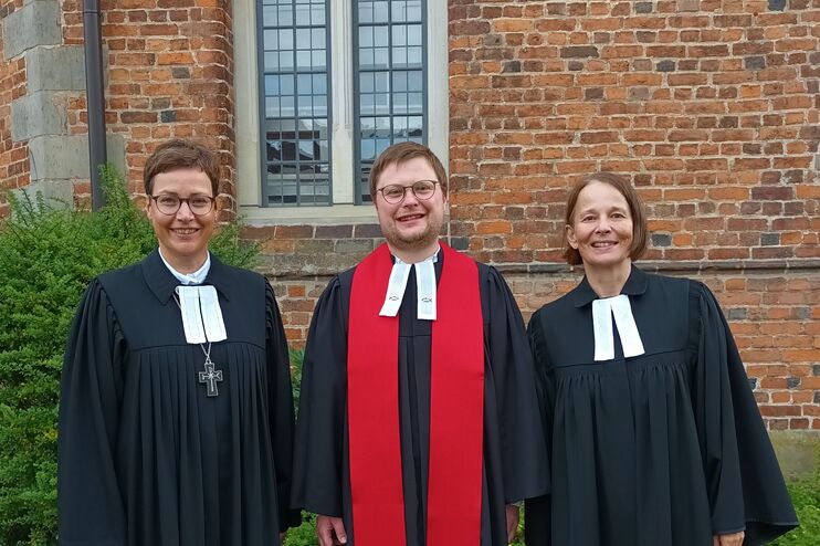 Zwei weiblich gelesene Personen und eine männlich gelesene stehen nebeneinander vor einer Backsteinkirche, alle tragen Talare. Die mittlere Person auch einen roten Schal.