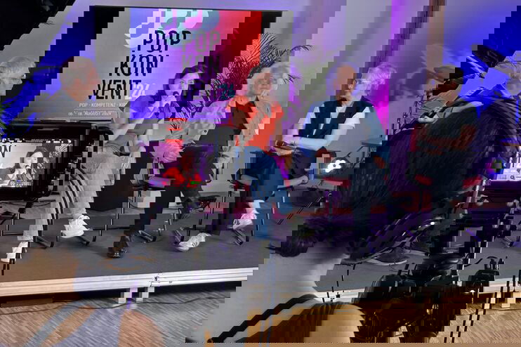 Zwei männlich gelesene und zwei weiblich gelesene Personen sitzen auf einem Podium, eine hält ein Mikro vor den Mund. Im Vordergrund ist jemand , der eine Kamera bedient. Im Hintergrund steht ein Bildschirm mit "popkonvent" auf rot-lila Hintergrund