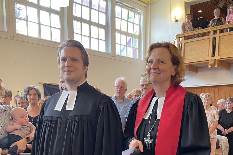 Eine männlich und eine weiblich gelesene Person, beiden in schwarzen Talare, gehen durch den Mittelgang einer Kirche zwischen stehenden Menschen hindurch.