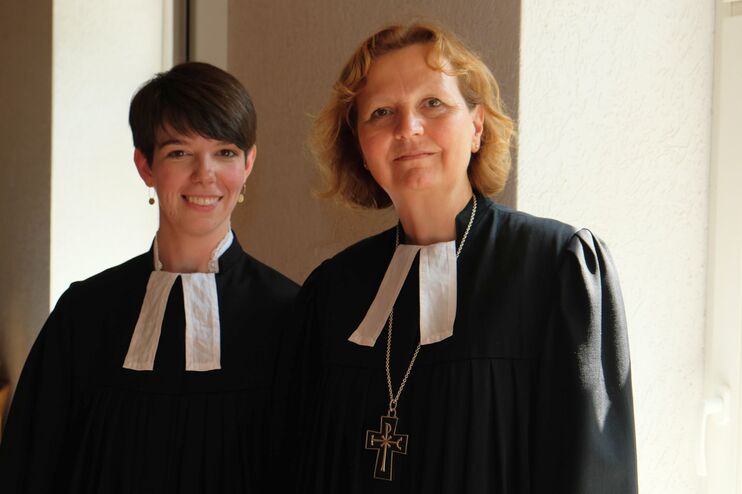 Zwei weiblich gelesene Personen mit Talaren stehen nebeneinander und lächeln.