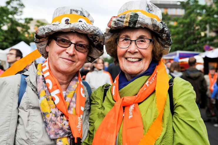 Zwei Frauen mit Hüten aus Papier und orangen Halstüchern auf einem belebten Platz