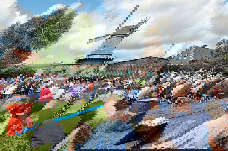 Auf einer Wiese sitzen viele Menschen auf Bänken und blicken nach vorn. Im Hintergrund eine Windmühle vor blauem Himmel mit weißen Wolken.