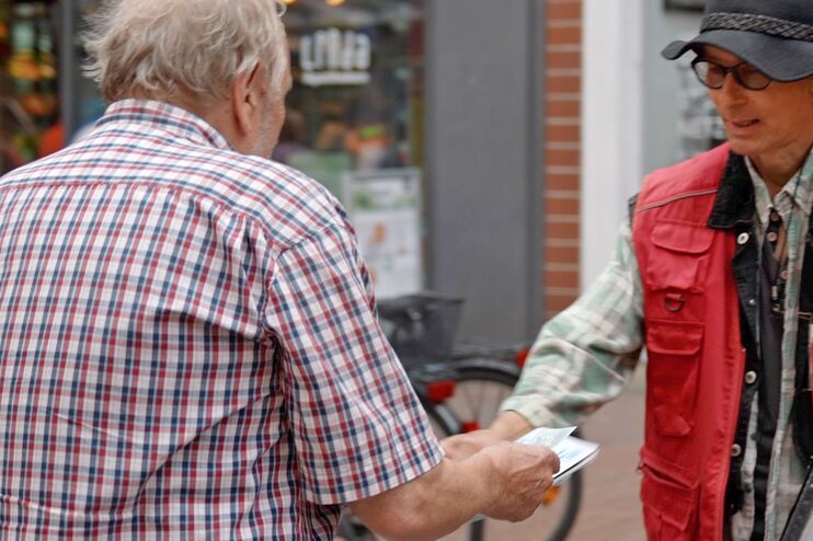 Zwei männlich gelesene Personen. Eine trägt eine rote Weste, auf der Asphalt. Die Person reicht der anderen eine Zeitung.