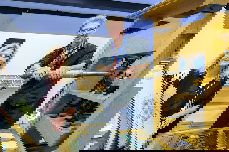 Eine als Frau und eine als Mann lesbare Person zeigen in einem Museum eine historische Druckmaschine. 