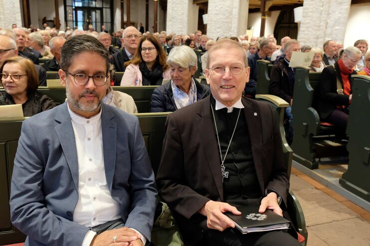 Zwei männlich gelesene Personen sitzen nebeneinander in einer Kirchenbank, viele andere Menschen ringsum. 