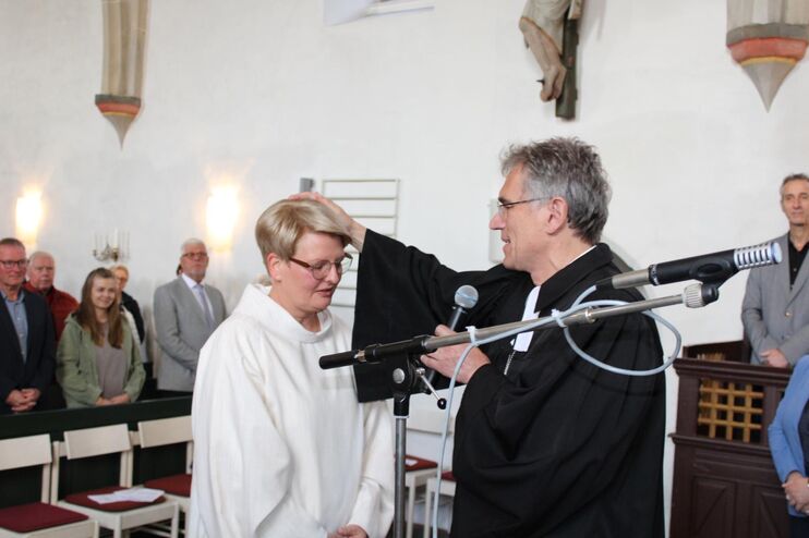 Eine weiblich gelesene Person in weißem Talar bekommt eine Hand von einer männlich gelesenen Person in schwarzem Talar auf den Kopf gelegt. 