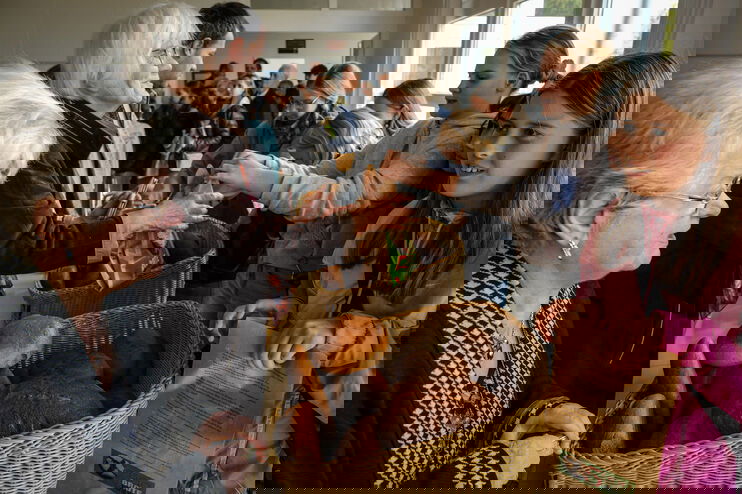 Jüngere Menschen stehen rechte von Brotkörben und bieten sie offenbar zum Kauf an. Links stehen ältere Menschen, eine Person nimmt gerade ein Brot entgegen.