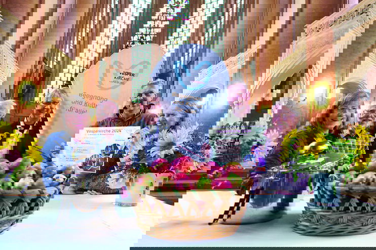 Blick in eine hohe Kirche, im Vordergrund ein gedeckter Tisch mit einer Schale Äpfel, Blumen und einem Kochtopf. Dahinter stehen fünf Personen und eine Flagge, auf der steht "Vesperkirche".