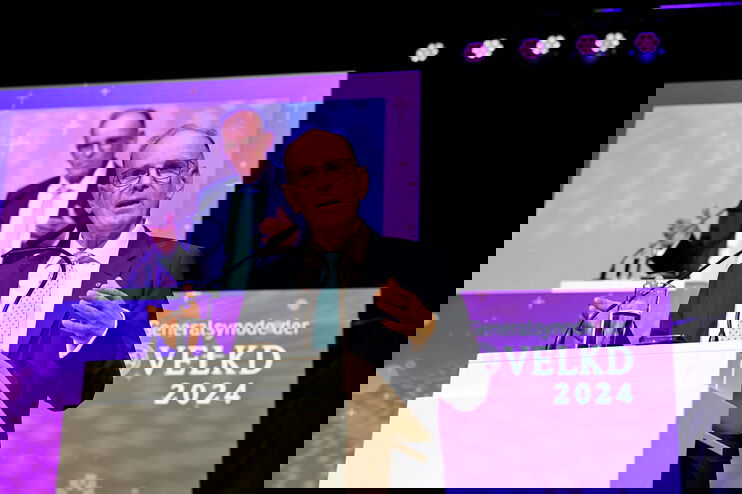 Eine als Mann lesbare Person steht an einem Redepult und spricht gestenreich. Hinter ihm ist er auf einer Leinwand zu sehen.