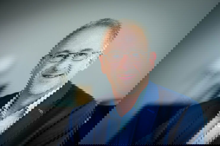 Eine als männlich lesbare Person im hellblauen Hemd, mit dunkelblauem Hemd und Brille vor grünem Hintergrund.