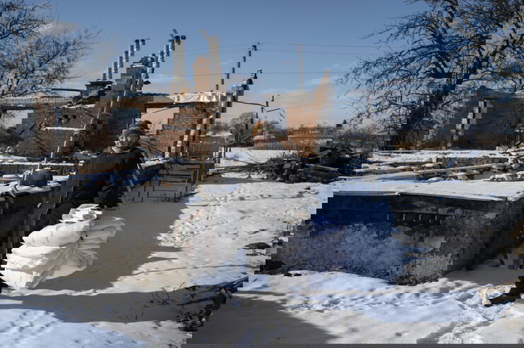 Eine Person steht mit zusammengebundenen Decken im Schnee zwischen Ruinen.