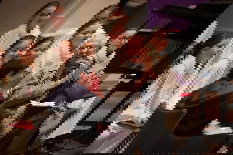 Zwei Personen, eine weiblich und eine männlich gelesene, sitzen nebeneinander, sich leicht zugewandt. Im Hintergrund junge weibliche Personen in weißen Gewändern.