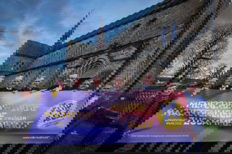 Eine weiblich zu lesende Person und sieben männlich zu lesende Personen halten ein Banner. Im Hintergrund ist eine Kirche zu sehen.