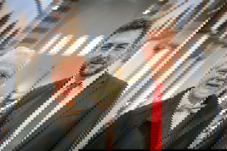 Zwei männlich gelesene Personen, eine älter mit weißem Haar und Brille und Fliege, neben einer jüngeren mit rotem Schlips in einer Kirche.