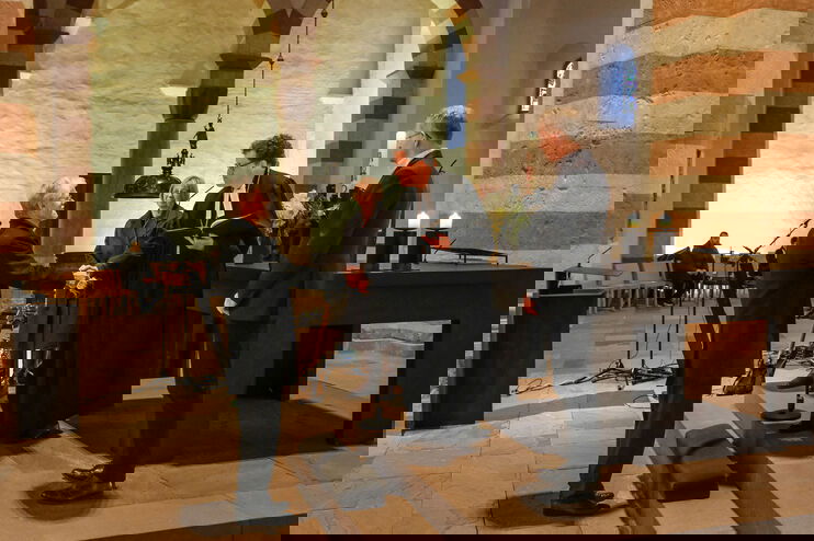 Drei Menschen stehen erhöht vor einer anderen Person. Die mittlere trägt Talar und reicht der entgegenstehenden Person die Hand.