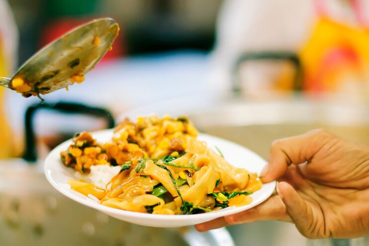 Ein Teller mit Essen, eine Person tut einer anderen mit einer Kelle was auf.