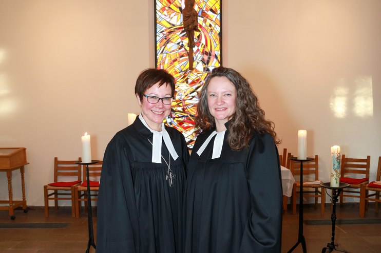 Zwei weiblich gelesene Personen in Talaren stehen vor einem Buntglasfenster mit Kreuz.