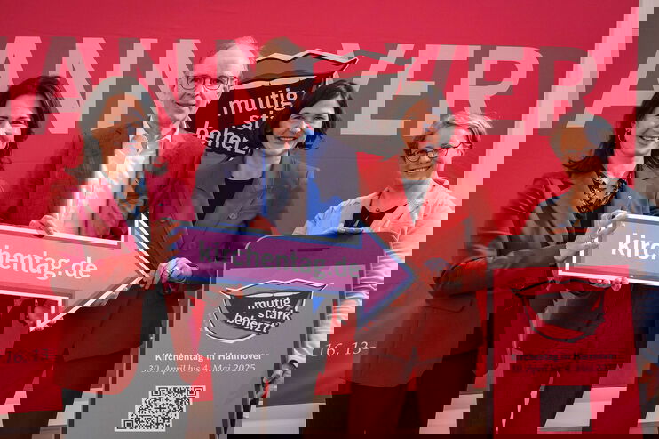 Drei als Frauen lesbare Personen und eine als Mann lesbare Person halten einen Pfeil mit der Aufschrift kirchentag.de und zeigen auf ein übergroßes Smartphone-Modell.