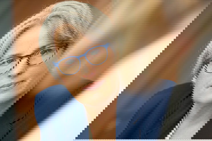 Eine als Frau lesbare Person mit blauer Brille und blauem Jacket hört einer anderen Person zu.