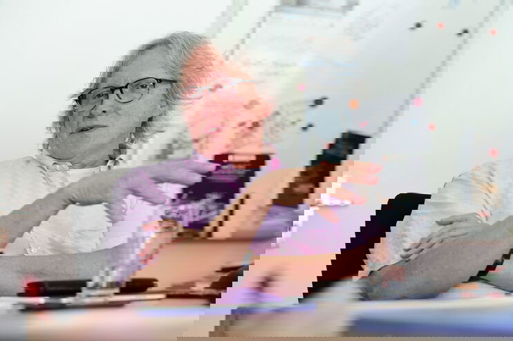 Eine als Mann lesbare Person im rosa Hemd sitzt an einem Tisch hinter einem Wasserglas und spricht.