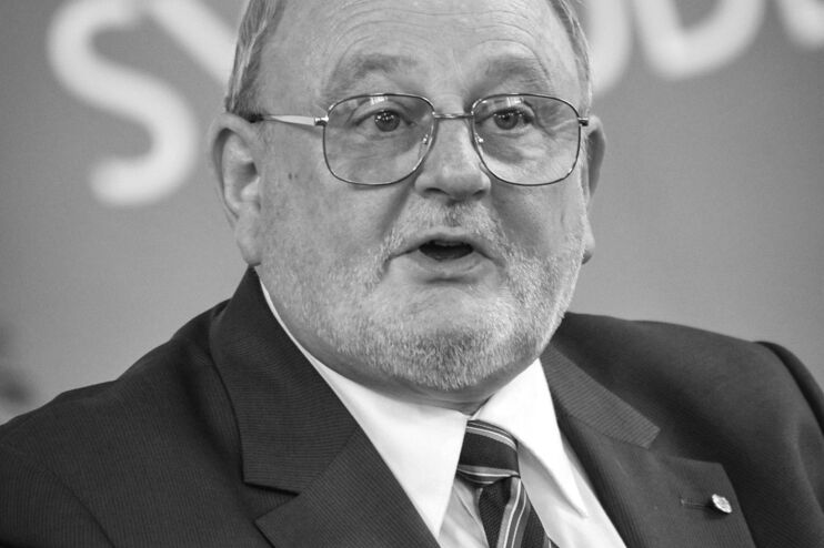 Foto einer als Mann lesbaren Person mit Brille und hoher Stirn hinter einem Mikrofon. Im Hintergrund steht Landessynode auf einem Banner.