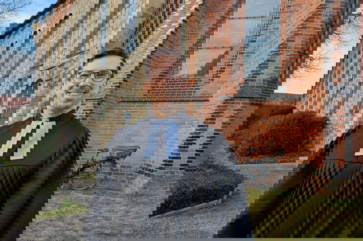 Eine als Mann lesbare Person im Talar steht vor einer Backsteinkirche.