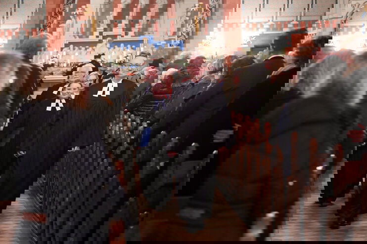 Eine männlich und eine weiblich gelesene Person, jeweils im Talar, gehen den Mittelgang einer vollbesetzten Kirche hinunter.