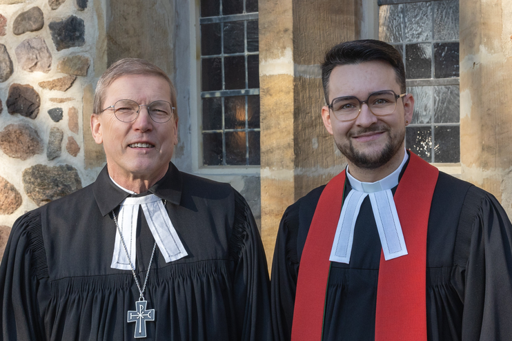 Zwei männlich gelesene Personen in Talaren stehen nebeneinander vor einer Steinwand, einer Kirche.