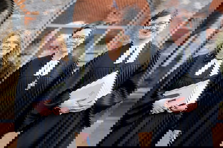 Zwei als Frauen lesbare Person und eine als Mann lesbare Person im Talar lächeln in die Kamera.