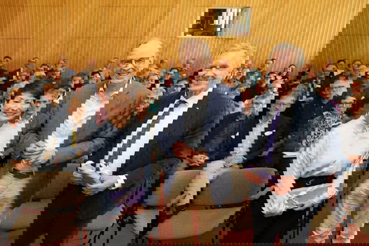 Eine als Frau lesbare Person und zwei als Männer lesbare Personen stehen vor einer größeren Gruppe in einem Tagungssaal.