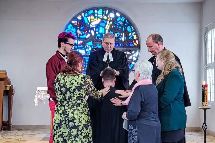 Sechs Personen, einer in Talar, stehen ringsum eine knieende Person. Sie befinden sich in einem Altarraum mit einem blauen Buntglasfenster im Hintergrund.
